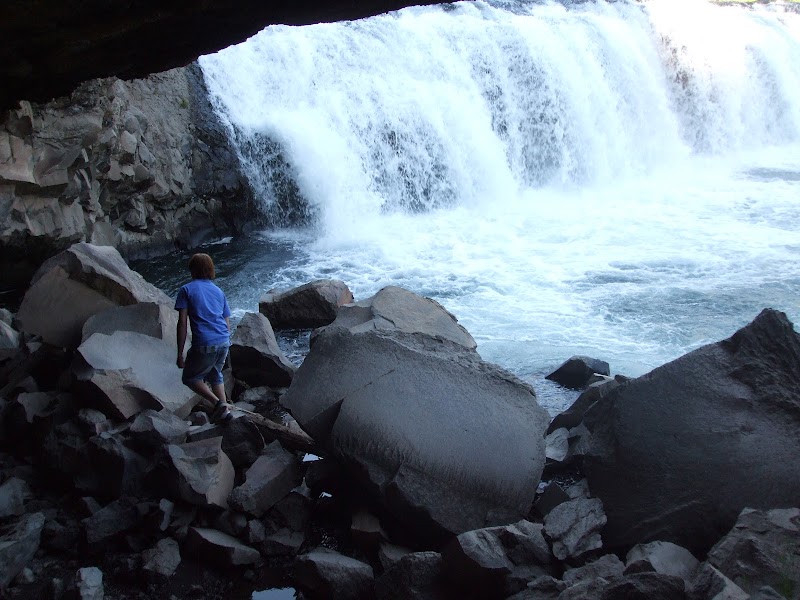 Person in a blue shirt walks among jagged gray rocks beside a tall white waterfall at Cave Falls, Yellowstone National Park.