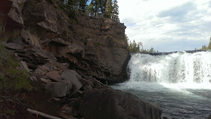 Cave Falls spills over a rocky ledge into a forested pool, framed by sheer cliffs, boulders, and evergreens in Yellowstone National Park.