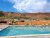 Poolside view with turquoise pool, yellow and green lounge chairs under blue umbrellas, orange wall, and red rock Arches National Park beyond.