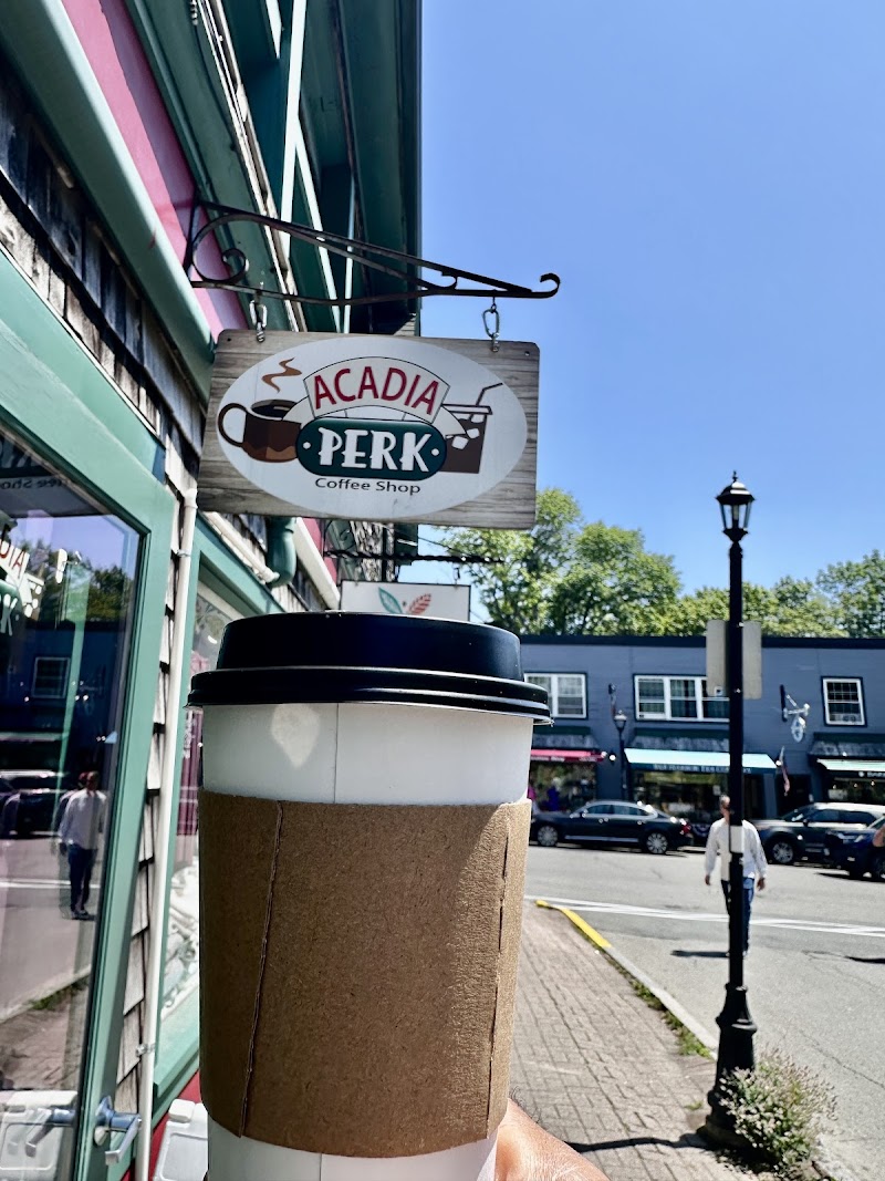 Sunlit street scene in Acadia National Park with a to-go coffee cup in the foreground and a cafe sign on a green storefront.