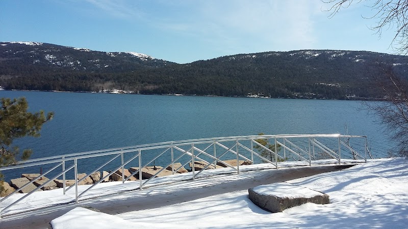 Snowy shoreline along a calm blue lake with a white railing, rocky steps, and forested hills in Acadia National Park.
