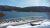 Snowy shoreline with a metal railing overlooks a calm blue lake and forested hills at Acadia National Park.