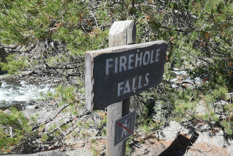 Wooden sign reading Firehole Falls sits beside a rocky, rushing stream with green pines in Yellowstone National Park.