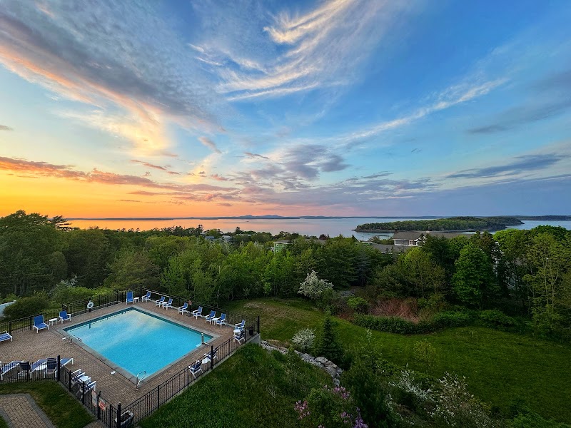 Sunset over the Acadia National Park coastline with a rectangular pool, deck seating, and lush trees.