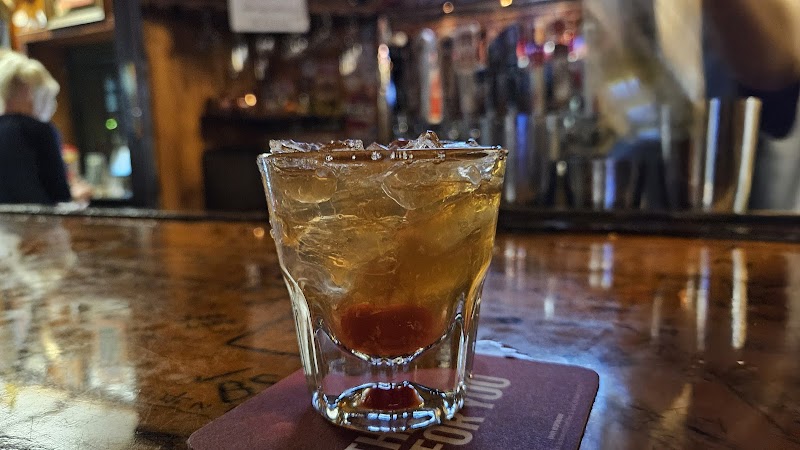 Whiskey on the rocks in a short glass rests on a wooden bar with ice and a cherry, inside Arches National Park tavern.