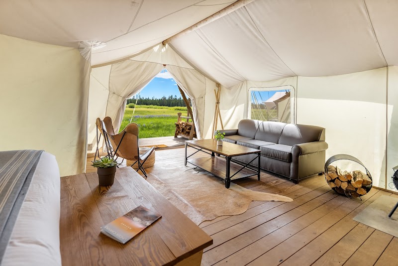 Lodge interior with a gray sofa, wooden coffee table, a firewood nook, and an open doorway to a grassy meadow in Yellowstone National Park.