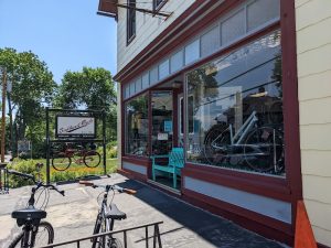 Sunlit storefront with large windows displaying bicycles, turquoise bench outside, and two bikes parked along a street in Acadia National Park.