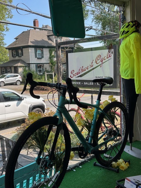 Teal road bike on a display stand in a shop window with a bright yellow helmet, overlooking a street near Acadia National Park.