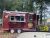 Maroon burger trailer operates as a park food stand with a menu board and colorful flowers nearby.