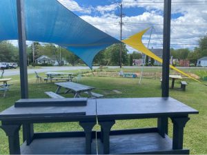In Acadia National Park, blue and yellow shade sails hover over wooden picnic tables on a grassy yard with benches and a road in the background.