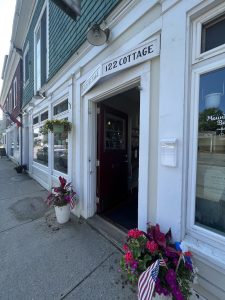 Open storefront with white facade, '122 Cottage' sign, hanging planters and bright flowers along Acadia National Park street.