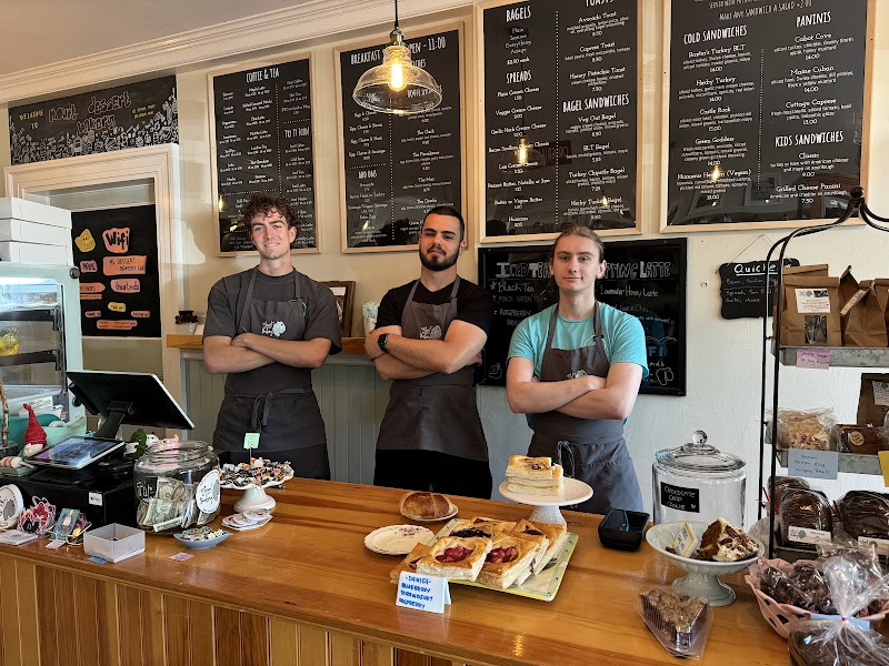 Three staff members in aprons stand behind a wooden counter with pastries, jars, and chalkboard menus in Acadia National Park.