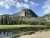Towering rocky dome rises above a dense pine forest, with a calm river and green meadow in Yellowstone National Park.