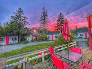 Patio deck with red chairs and a glass table under a closed umbrella at dusk, pink sunset over cottages in Acadia National Park.