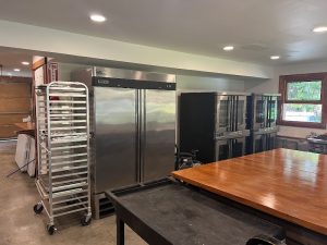 Commercial kitchen with stainless steel refrigerator, rolling rack, prep table, ovens near a window in Acadia National Park.