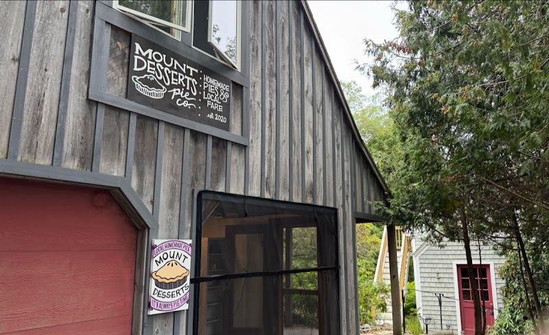 Rustic gray wooden storefront with a chalkboard sign, a large front window, and a pink door beside trees in Acadia National Park.