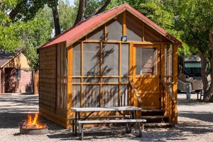Wooden cabin with a red pitched roof and screened front, a picnic table and fire pit in a shaded Arches National Park campground.