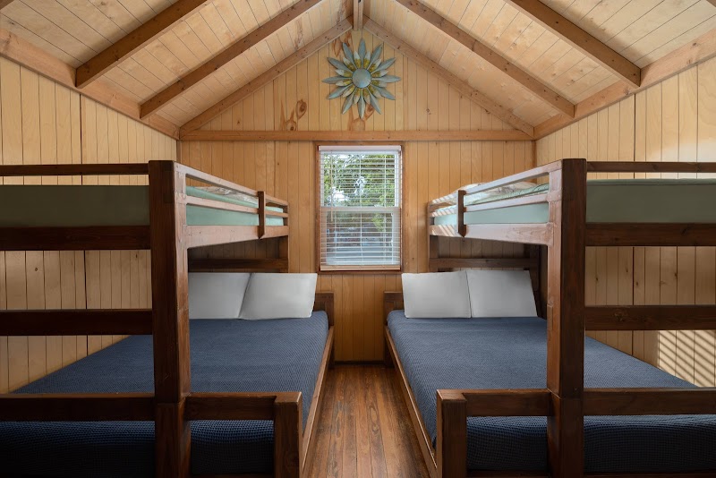 Inside a wooden cabin, two sets of sturdy bunk beds with blue mattresses face a central window, sun decoration above, in Arches National Park.