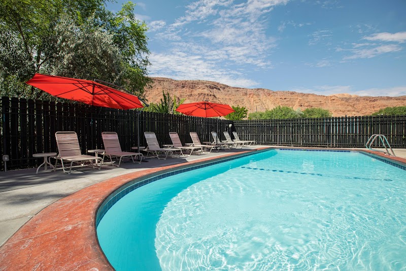 Curved blue pool with red umbrellas and lounge chairs along a fenced deck; desert hills loom in Arches National Park.