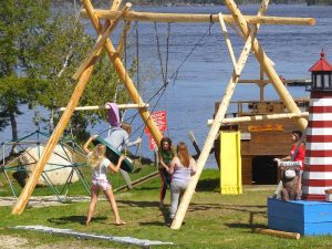 Balsam Cove Campground at Acadia National Park features a wooden swing set near the shoreline with kids playing in a sunny campsite.