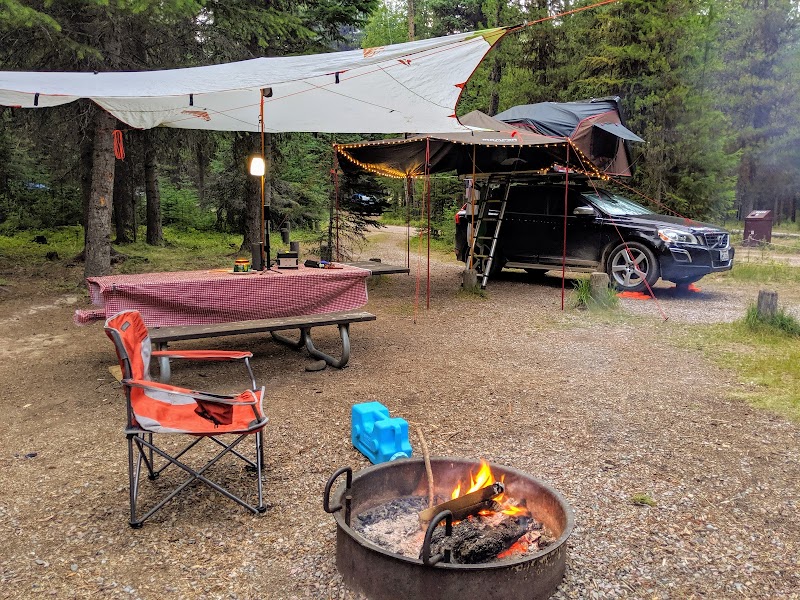 Glacier National Park campground with a black SUV under a tarp shade, string lights, picnic table, chairs, and a fire pit.