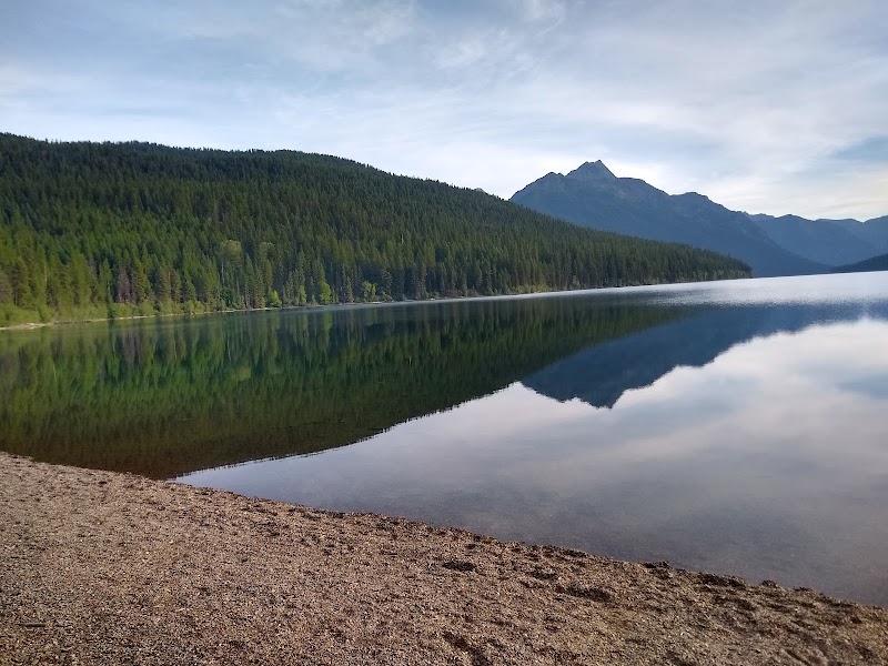 Glacier National Park: a calm lake with evergreen shoreline, pebbled beach, and mountains reflected on the glassy water.