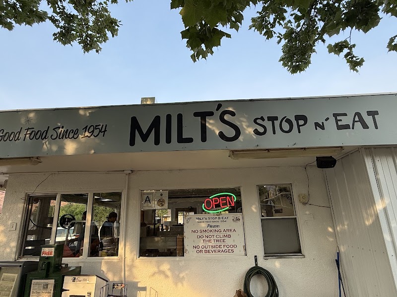 A small roadside diner storefront near a shade-dappled entrance in Arches National Park on a sunny day, inviting visitors to stop for a quick meal.