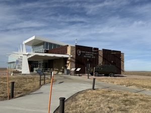 Modern visitor center with glass walls and a white angular sculpture, stone facade, and a green armored vehicle at Badlands National Park.