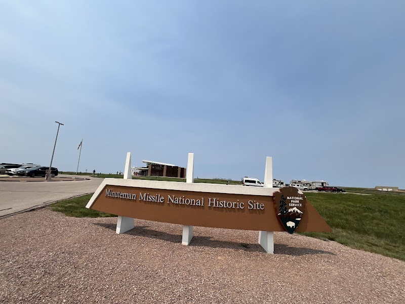 Bright day at Badlands National Park shows a brown angled sign for a historic missile site with several cars in parking lot.