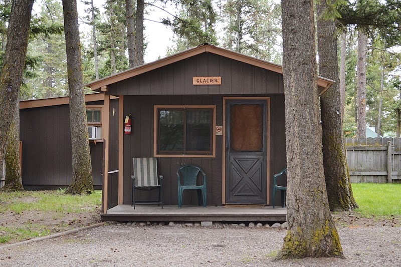 Small dark brown cabin with a raised porch and chairs, tucked among pines in Glacier National Park.