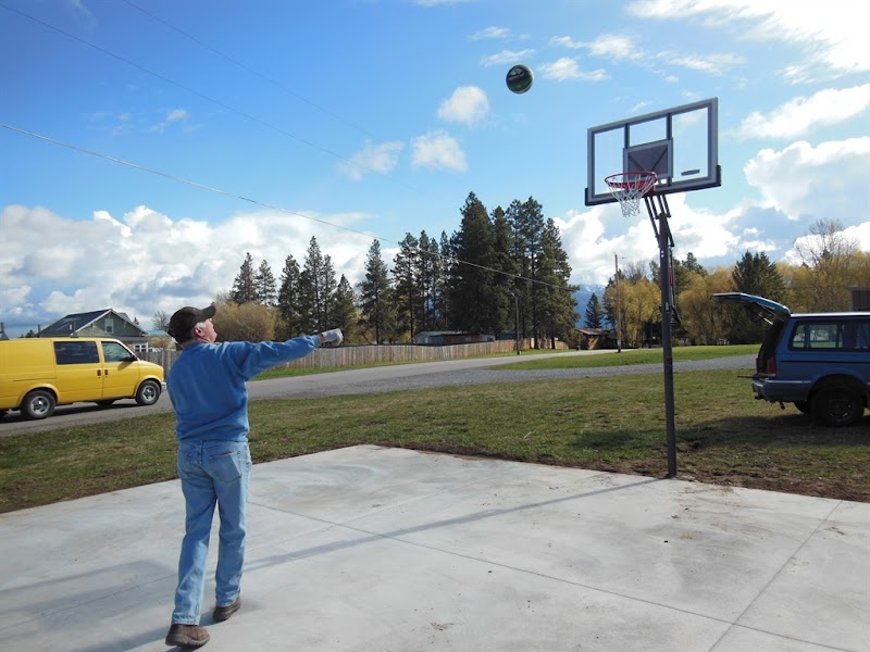 Man shooting a basketball on a concrete court with hoop, yellow van, blue SUV, and pine trees in Glacier National Park
