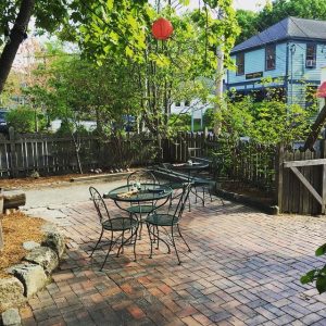 Brick-paved cafe courtyard with round metal tables and chairs, a red lantern, and green shrubs in Acadia National Park.