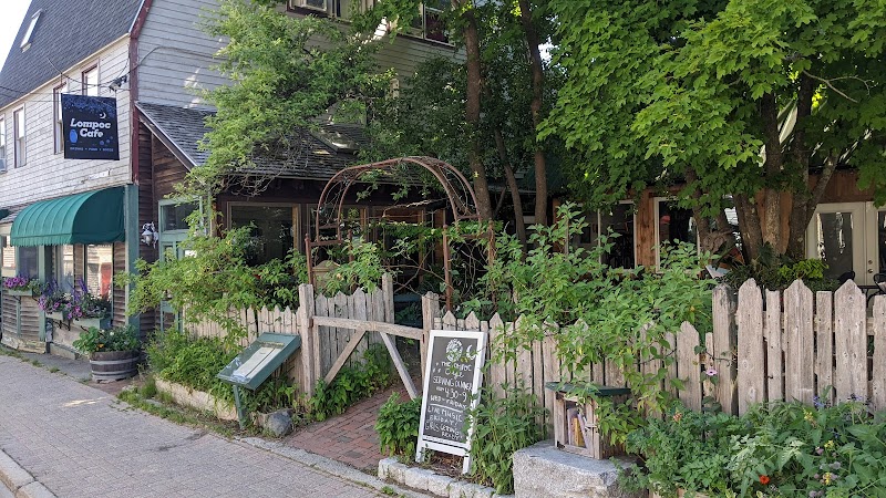 Cozy cafe storefront with a wooden picket fence, arch trellis, chalkboard menu, and lush greenery along a brick sidewalk in Acadia National Park.