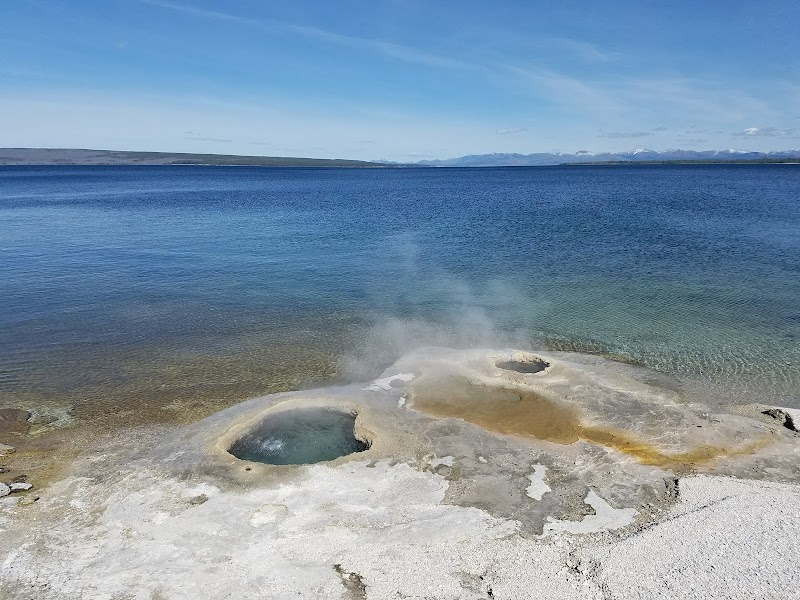 Yellowstone National Park: steaming mineral pools along a mineral-streaked shoreline beside a calm blue lake with distant mountains.