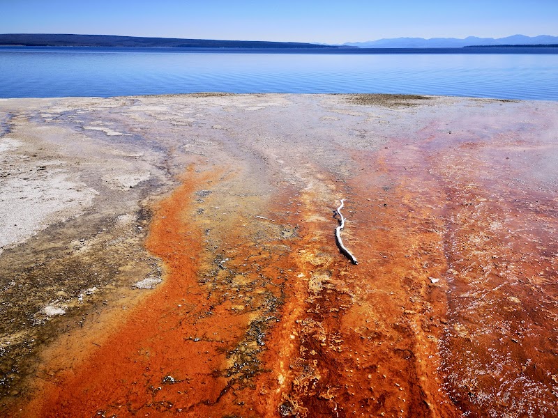 Bright orange and red mineral terraces extend toward a calm blue lake beyond, with distant mountains in Yellowstone National Park.