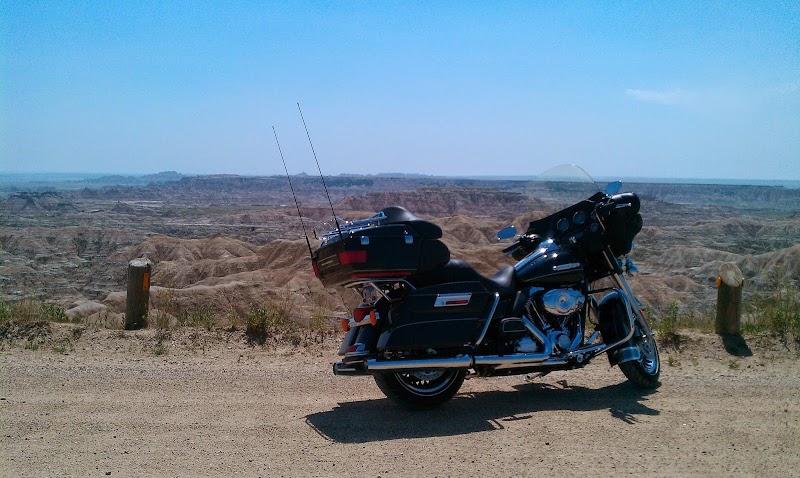 Black touring motorcycle with two fishing rods on the rear rack, parked on a dirt overlook in Badlands National Park with eroded canyon ridges in the distance.