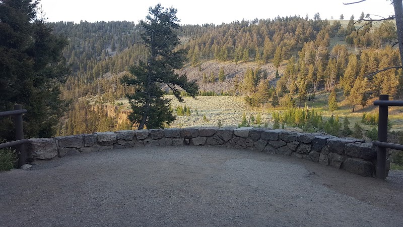 Overlook with a low stone wall and dirt railing, framing pine forest, rolling hills, and a meadow in Yellowstone National Park.