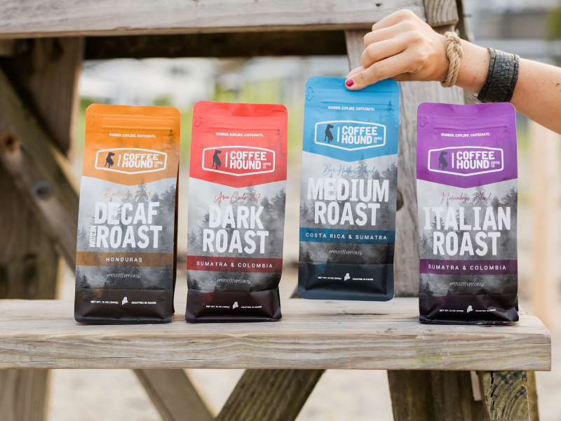 Row of four coffee bags in bright orange, red, blue, and purple on a wooden bench, a hand holds the blue bag in Acadia National Park.