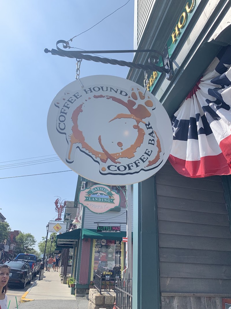 A circular coffee splash sign hangs from a bracket outside a green storefront with red-white-blue bunting in Acadia National Park.