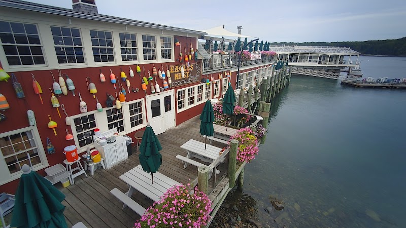 Red waterfront building adorned with colorful buoys, wooden deck with white tables and green umbrellas at Acadia National Park.