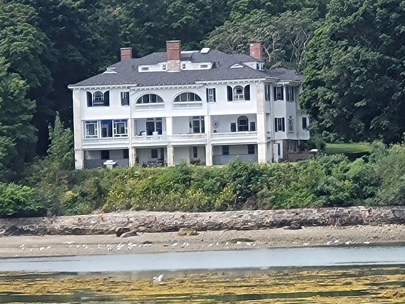 White, multi-level coastal house with arched windows and balconies sits among trees on a rocky shoreline near Acadia National Park.