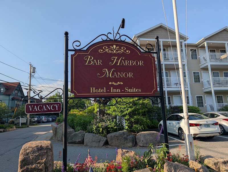 Lodging sign with maroon panel beside a multi-story hotel, cars parked, and flowering beds in Acadia National Park.