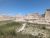 Dirt trail winds through grassy badlands toward layered sandstone cliffs under a blue sky in Badlands National Park.