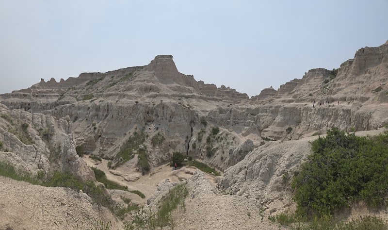 Dusty, eroded Badlands with a narrow trail descending into gullies and scattered shrubs at Badlands National Park.