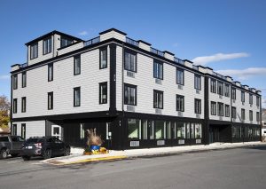 Modern gray three-story hotel with black trim sits on a street in Acadia National Park, with parked cars out front.