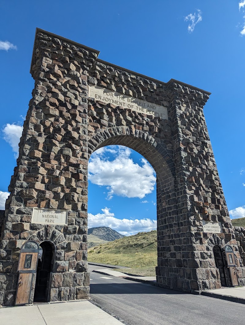 Massive stone Roosevelt Arch gateway at Yellowstone National Park with a blue sky, arched opening, and grassy hills beyond.