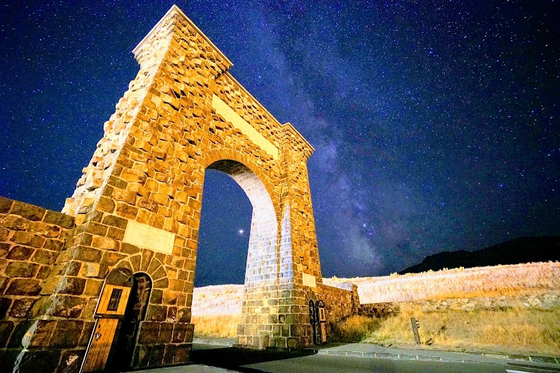 Stone arch gateway built from rough-cut stones stands over a road in Yellowstone National Park, beneath a starry night sky.