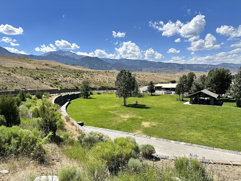 Green lawn in a Yellowstone National Park picnic area with a curved stone path, trees, a shelter, and distant mountains under a bright blue sky.
