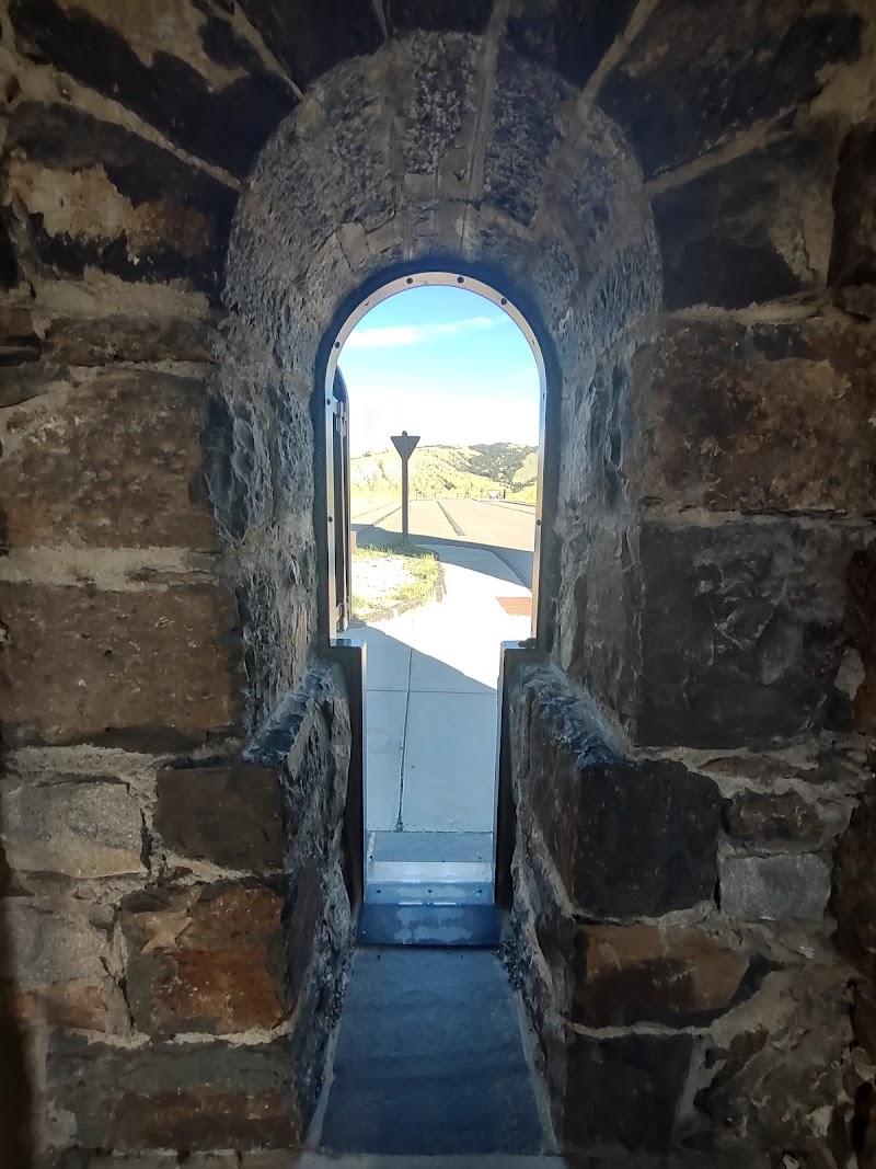 View through a rough stone archway opening to a paved path and road with distant hills under blue sky in Roosevelt Arch, Yellowstone National Park.