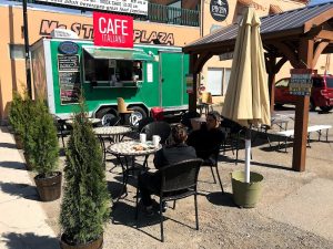 Outdoor seating area with round metal tables, black chairs, a beige umbrella, and a bright green mobile cafe trailer at Arches National Park.